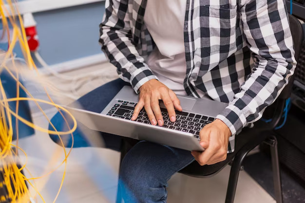 person writing in a notebook beside by an iPad, laptop, printed photos, spectacles, and a cup of coffee on a saucer