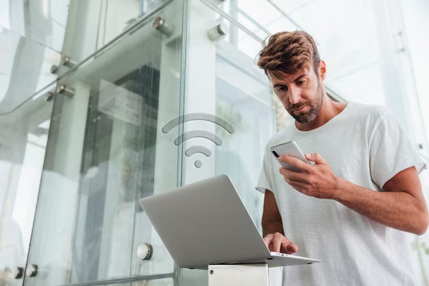 person writing in a notebook beside by an iPad, laptop, printed photos, spectacles, and a cup of coffee on a saucer